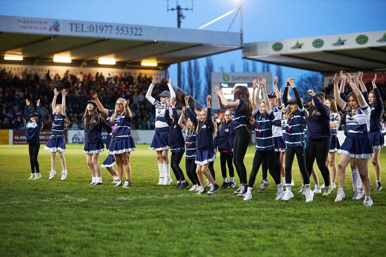 A group of girls dancing on a football pitch at a stadium