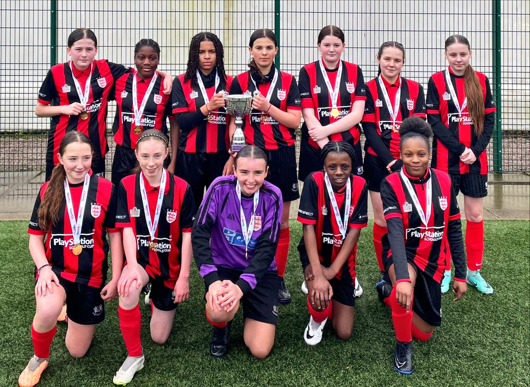 A football team of girls smiling for a photo. The girls all have medals on, and some are holding a trophy in the air. 