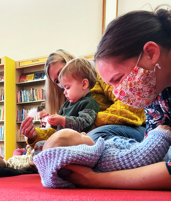 A mother holding a baby on the floor. A second mother playing with a baby on the back