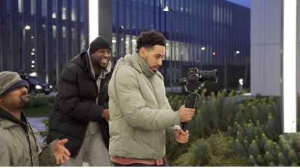 A man is operating a camera to film something out of shot. Two other men standing behind him are smiling. 