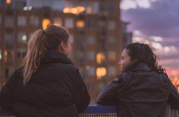 Two young women chatting on a balcony at dusk 941 x 641px