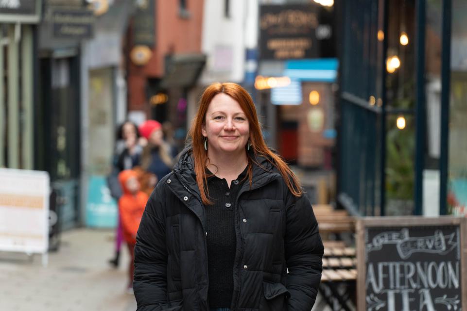 A white lady with red hair and a black coat stands in front of shops
