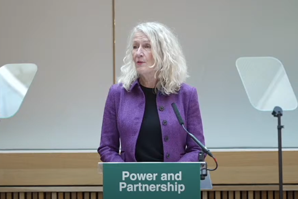 Louise Gittins wearing a purple jacket standing in front of a lectern speaking 