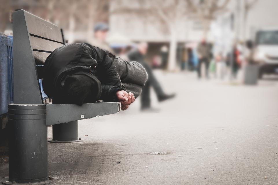 Homeless person asleep on a bench with people walking past