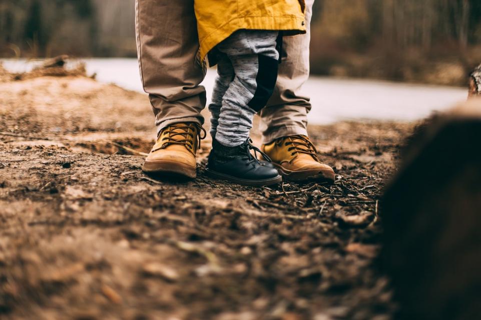 The legs of an adult in slacks and boots and a toddler in grey joggers, black boots and a yellow rain coat